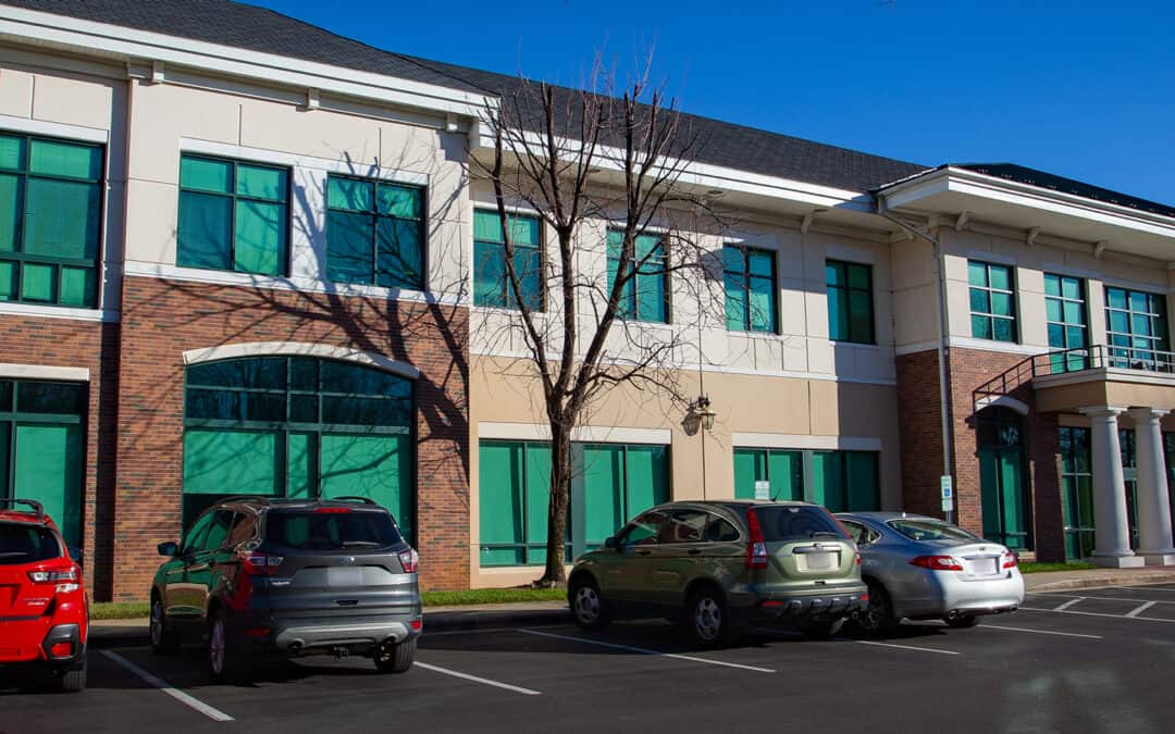 Exterior view of a brick and beige building with green-tinted windows and a parking lot with several cars.
