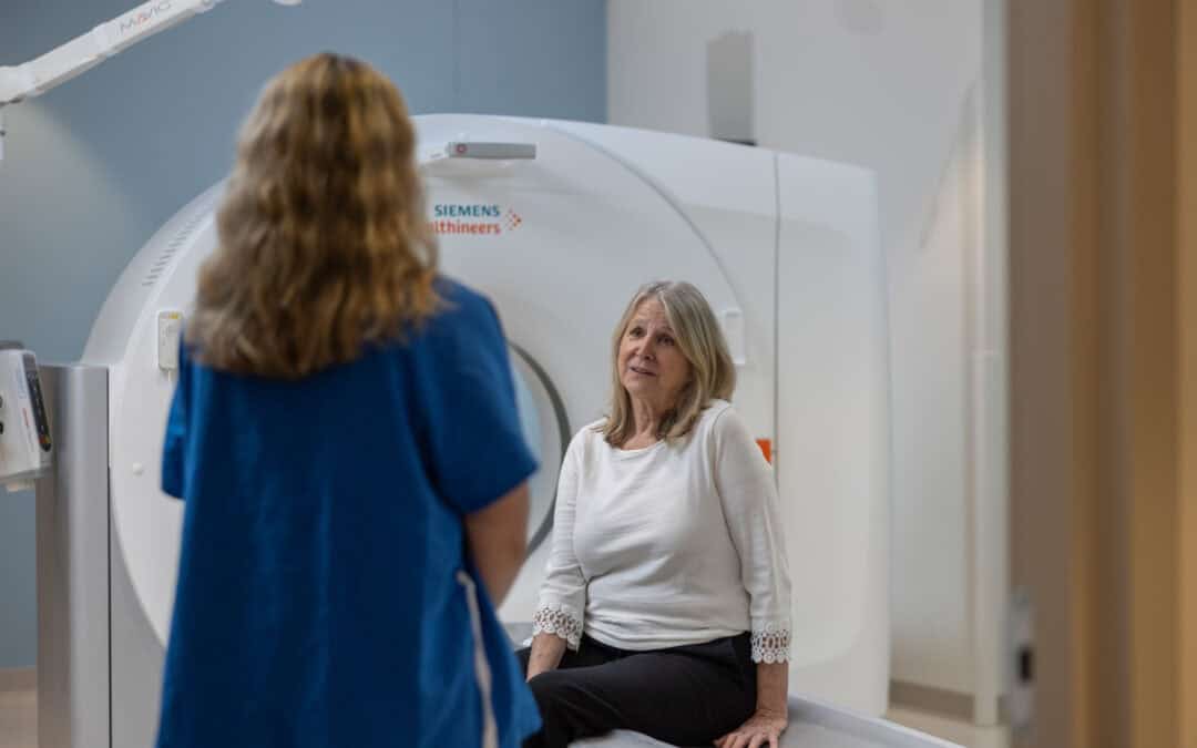 Female patient sitting on a Siemens Healthineers CT scanner table, talking to a medical professional.