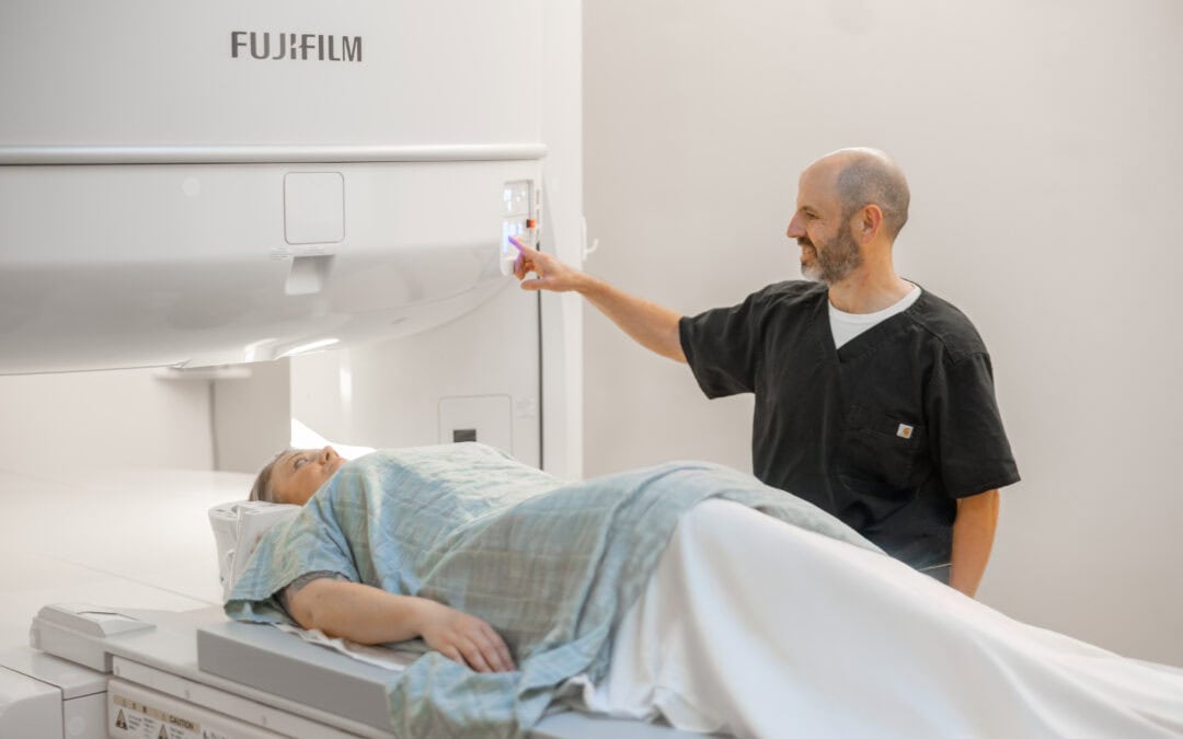 Male technician in black scrubs operating a Fujifilm imaging machine for a female patient lying on a table.